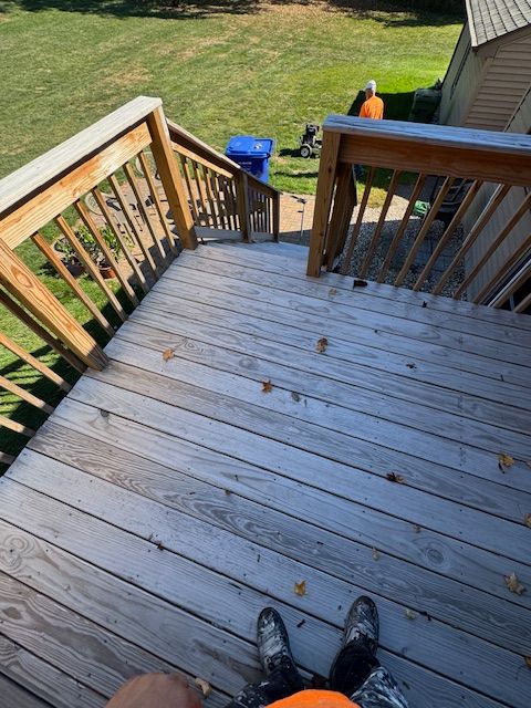 Wooden deck with steps leading down to yard. Person's feet visible. Blue trash can and person in orange shirt.