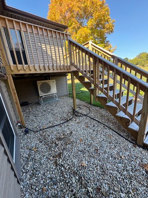 Wooden deck with stairs; gravel ground. Outdoor AC unit underneath. Green lawn and fall foliage in background.