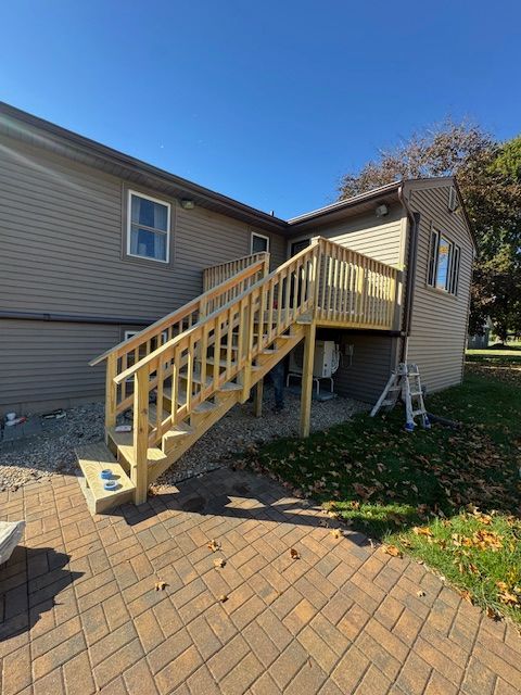 Wooden deck and stairs attached to a beige house, against a blue sky, on a brick patio.