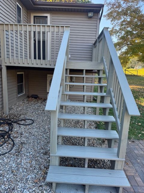 Exterior wooden stairs leading up to a deck, painted gray. Gravel and brick surround the stairs, with a grass lawn in the background.