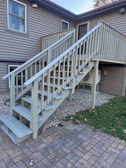 Wooden outdoor staircase leading up to a deck, painted light gray. Located next to a brick patio and a grassy area.