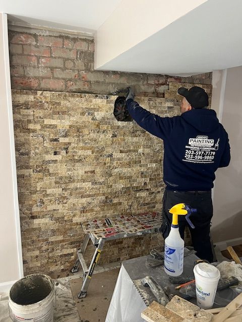Man installing stone tile on a wall, standing on a step stool. Tools and materials are nearby.