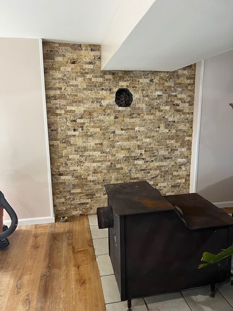 Dark metal wood stove in front of a brick wall with a vent and white trim. Wooden floor.