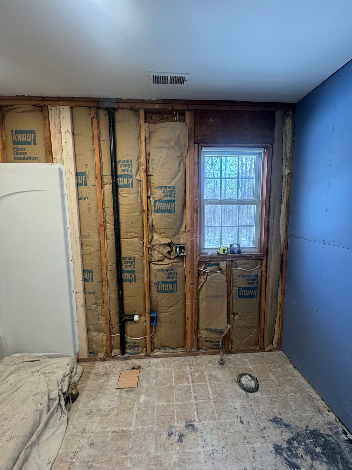 Bathroom renovation: exposed wall studs, insulation, window, blue wall, tub, and debris on floor.