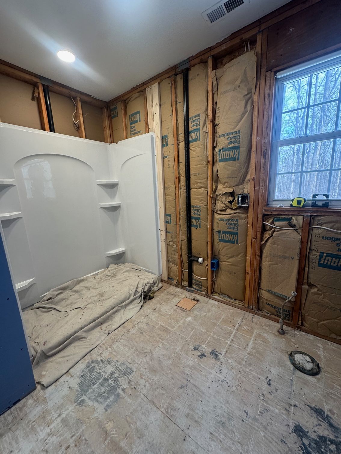 Bathroom under renovation: Exposed wall studs, insulation, white shower unit, window, and damaged floor.
