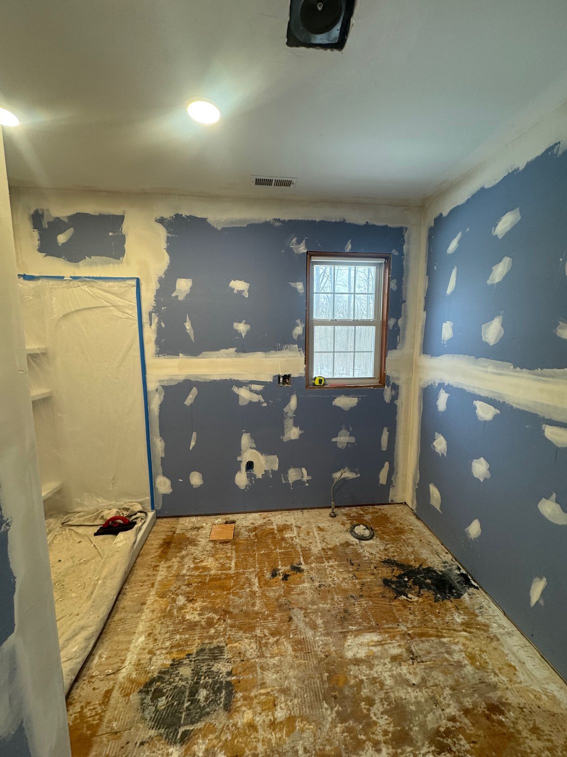 Bathroom renovation in progress, blue drywall, exposed subfloor, white shower stall, window, ceiling vent.
