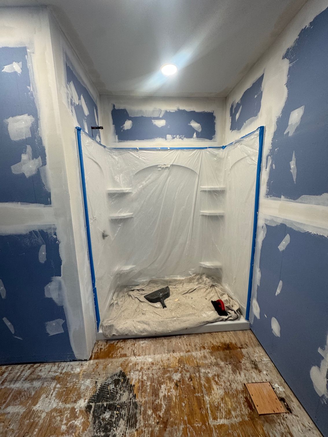 Bathroom under renovation with blue drywall, white shower stall covered with plastic, and tools on floor.