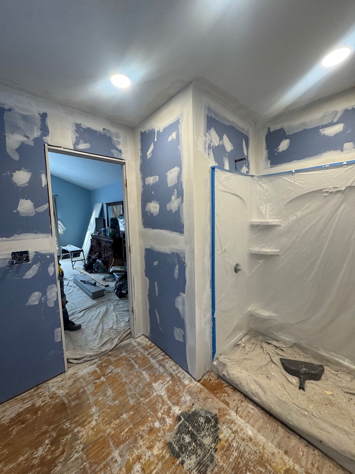 Bathroom renovation in progress. Blue drywall with spackle. Shower stall covered in plastic. Construction tools on the floor.