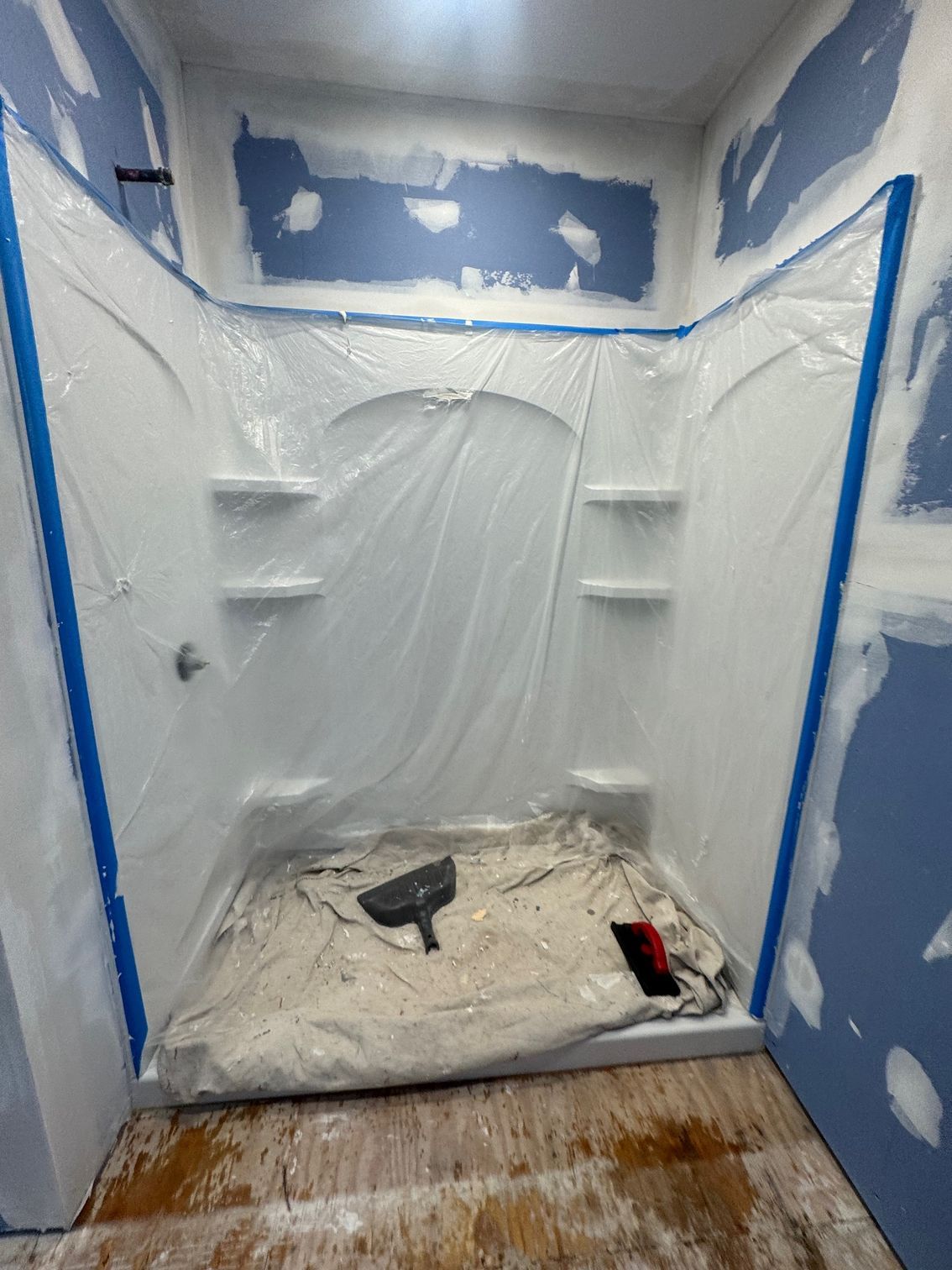 Shower stall covered in plastic sheeting for painting, blue tape edges, drywall on walls.