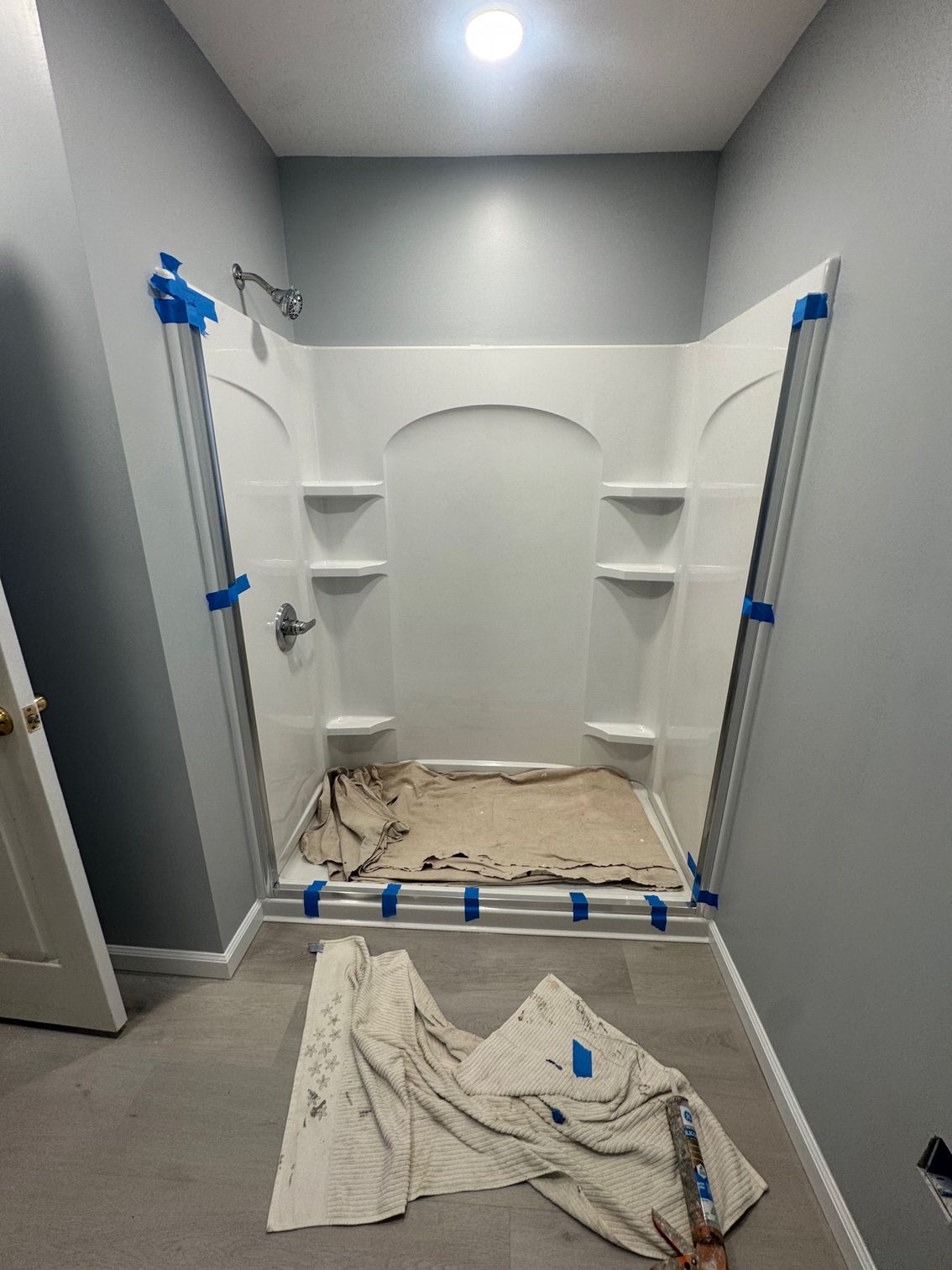 Empty white shower stall with built-in shelves, grey walls, and blue painter's tape; towel on floor.