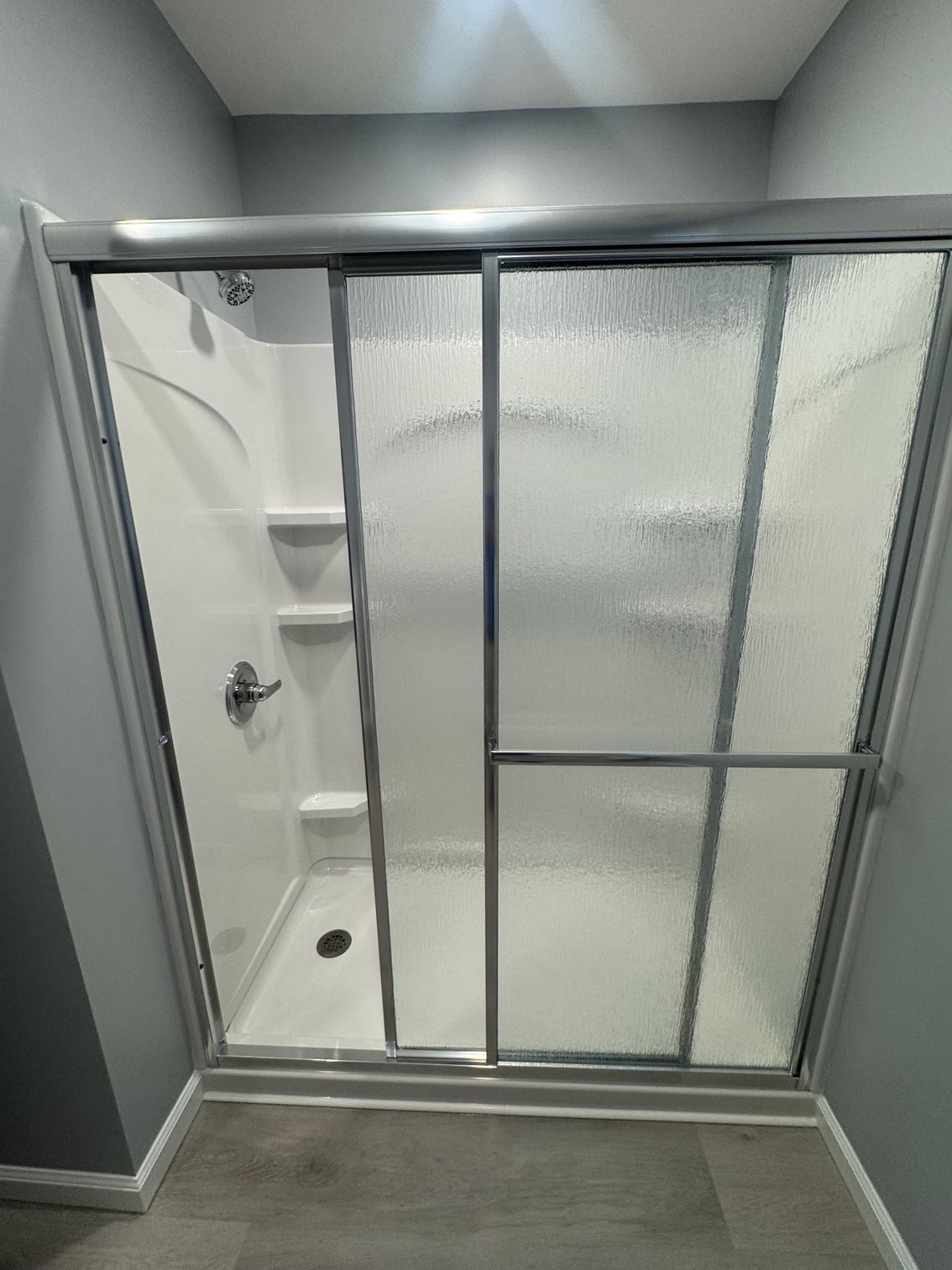 White shower stall with sliding glass doors, silver frame, three shelves, and a showerhead.