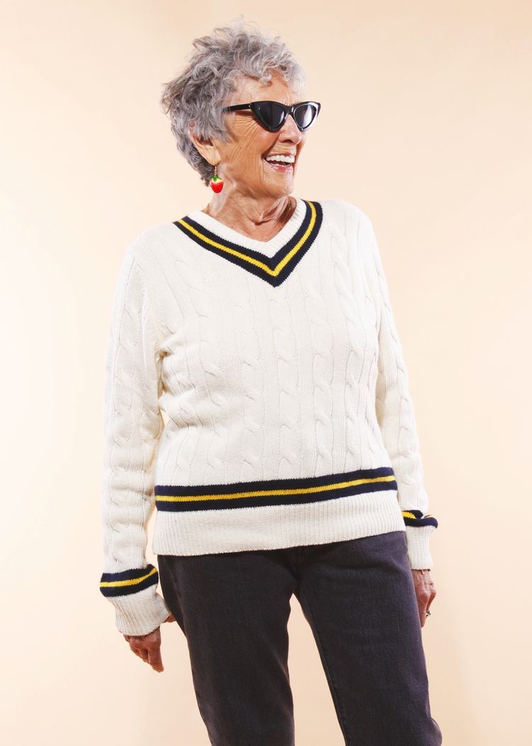 Woman wearing sunglasses and a cream-colored sweater smiles against a peach background.