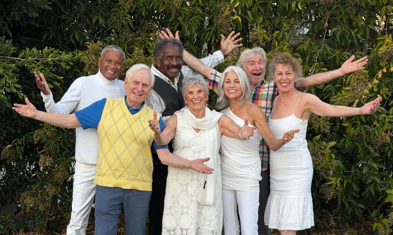 Group of people with arms outstretched, smiling in front of green foliage. White, blue, and yellow clothing.