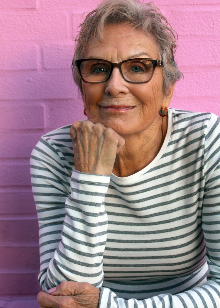 Woman with short gray hair and glasses rests chin on hand, striped shirt, pink brick wall.