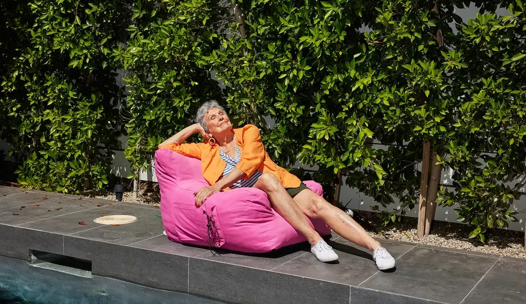 Woman in orange jacket reclines on pink beanbag by pool.