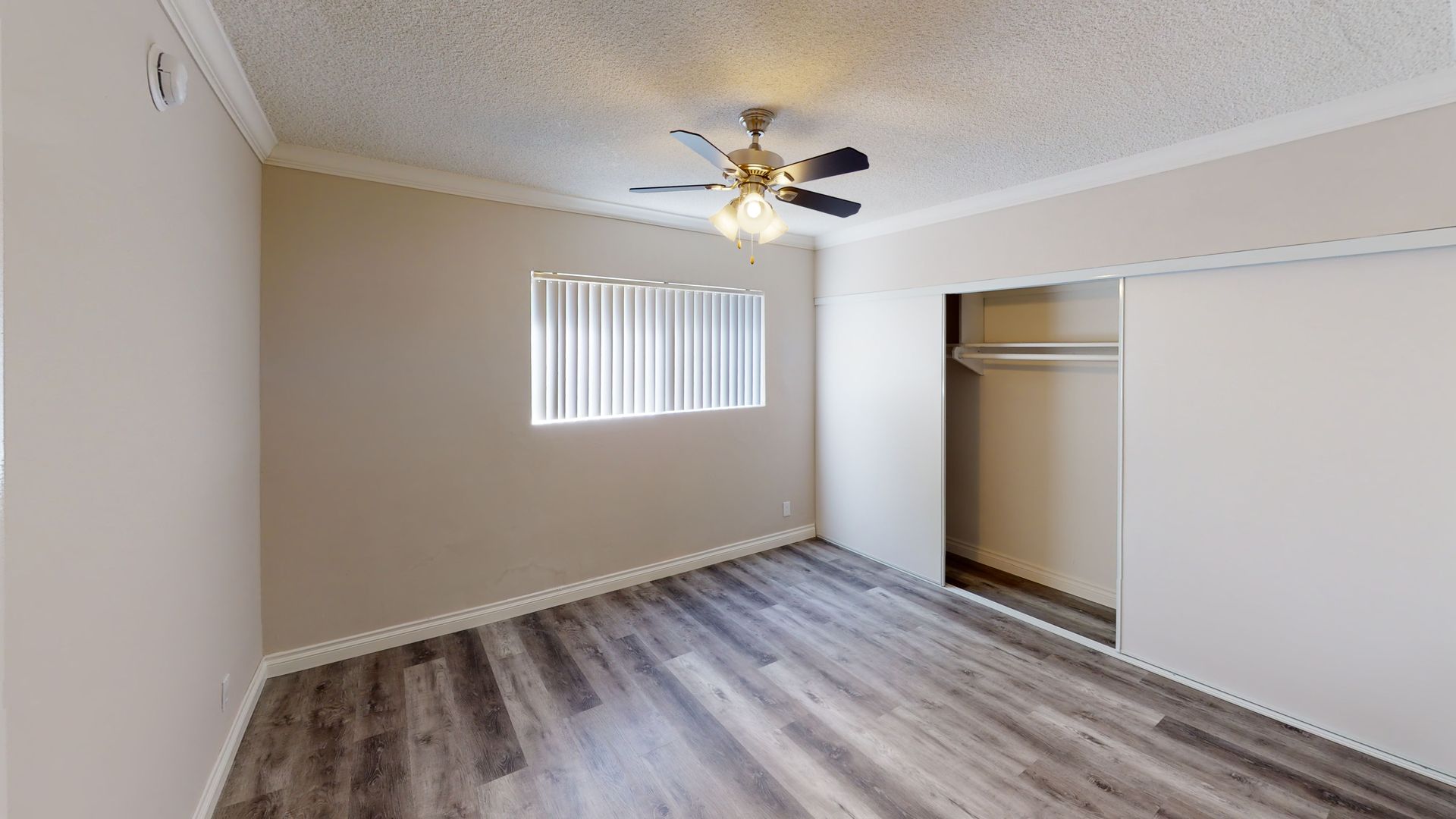 Empty bedroom with gray wood-look flooring, beige walls, sliding closet doors, and a ceiling fan.