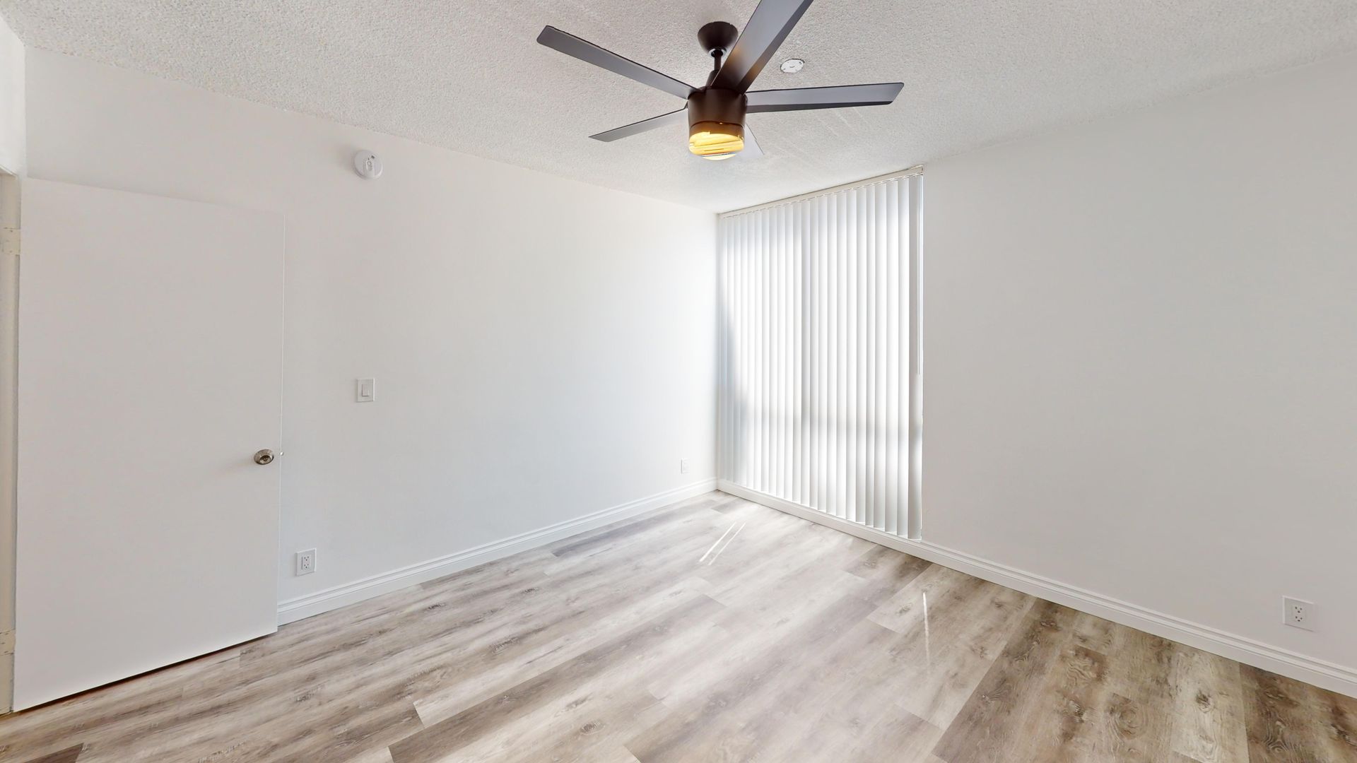 Empty, bright bedroom with a ceiling fan, window with vertical blinds, and light wood flooring.
