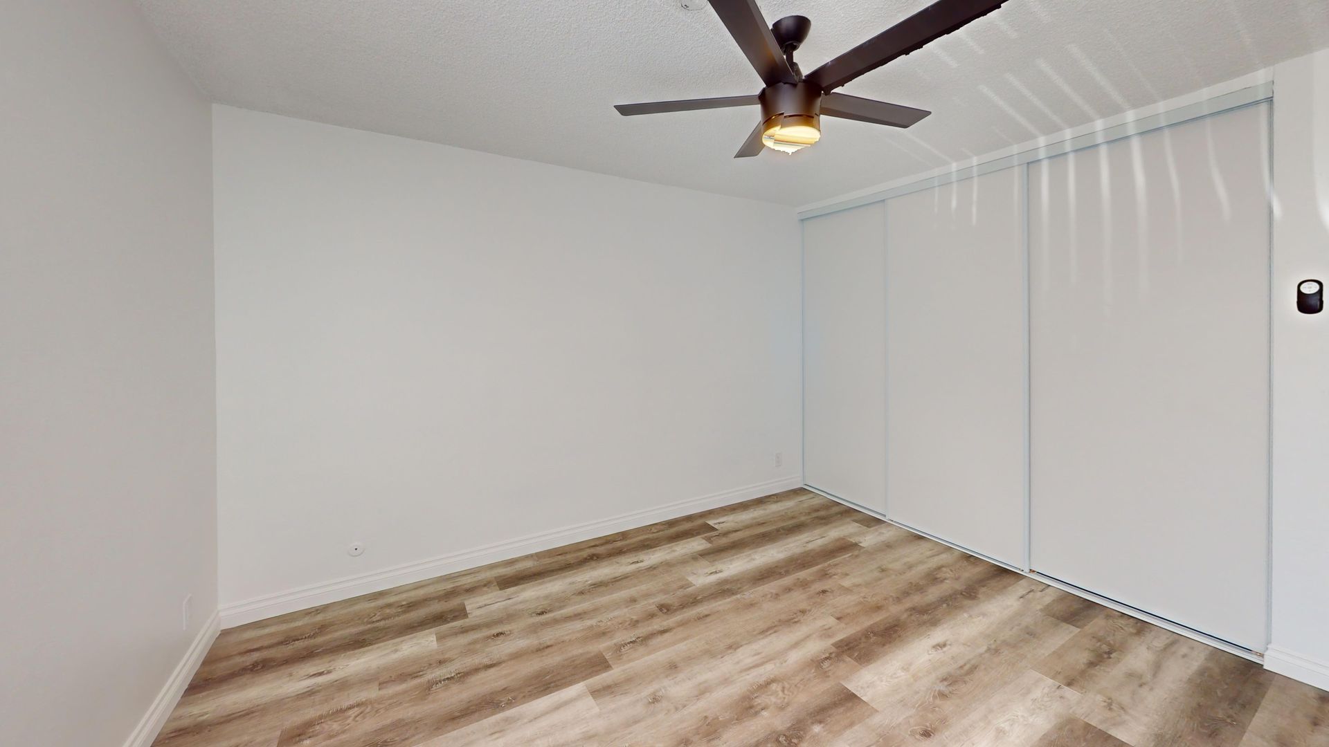 Empty room with wood-look floor, white walls, ceiling fan, and sliding closet doors.