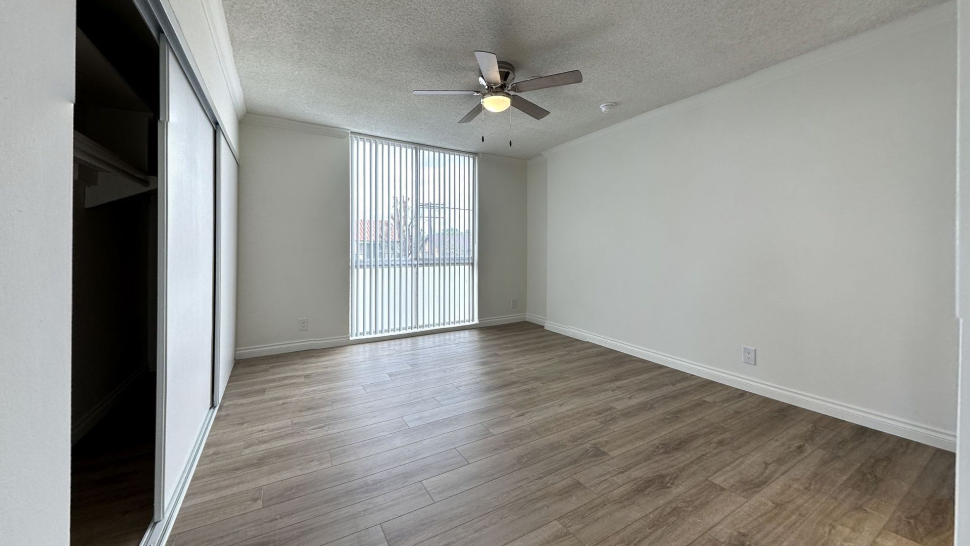 Empty bedroom with wood-look floor, large window with vertical blinds, closet, ceiling fan, and white walls.