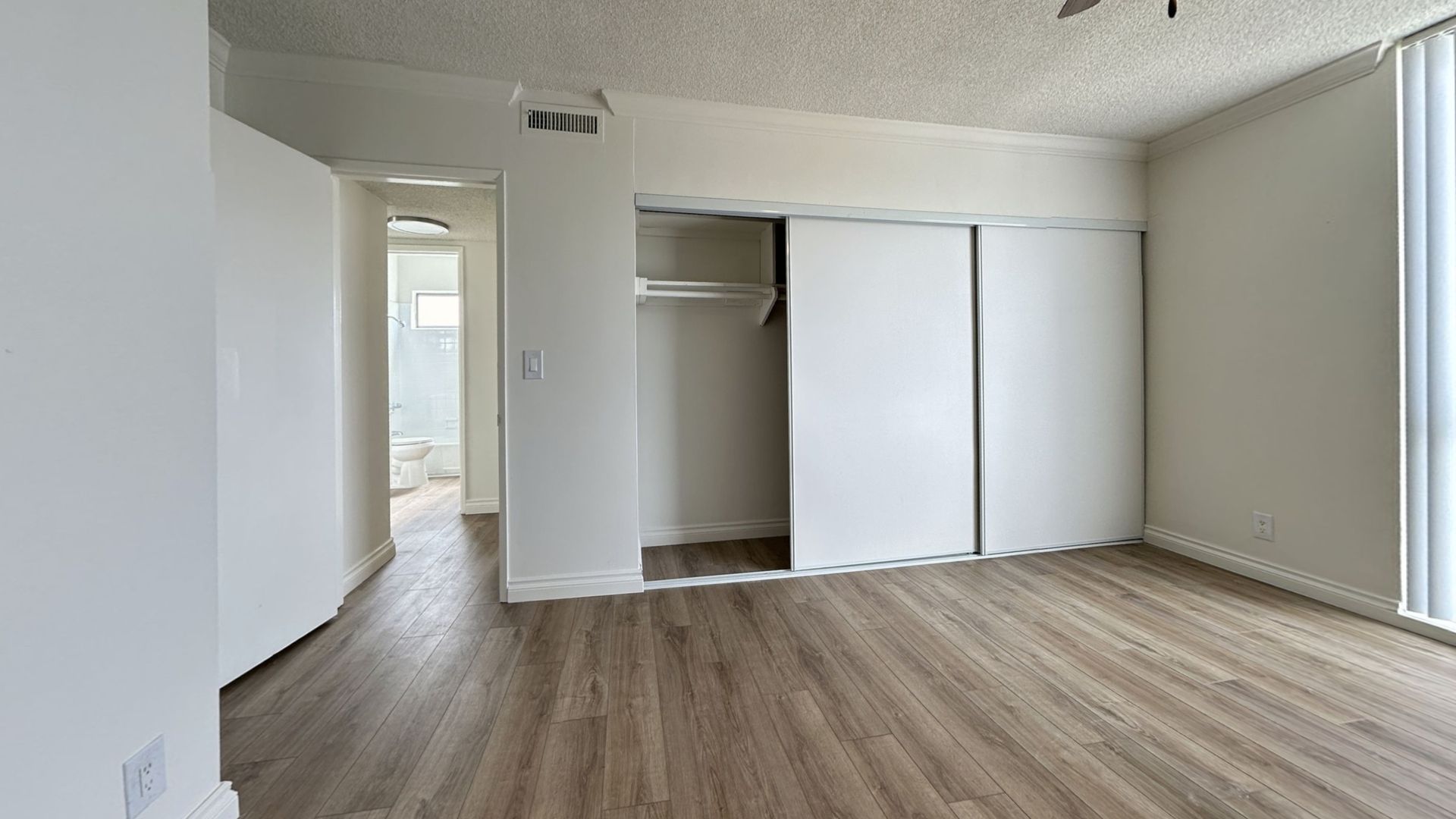 Empty room with white walls, sliding closet doors, and wood-look flooring. Hallway leads to a bathroom.
