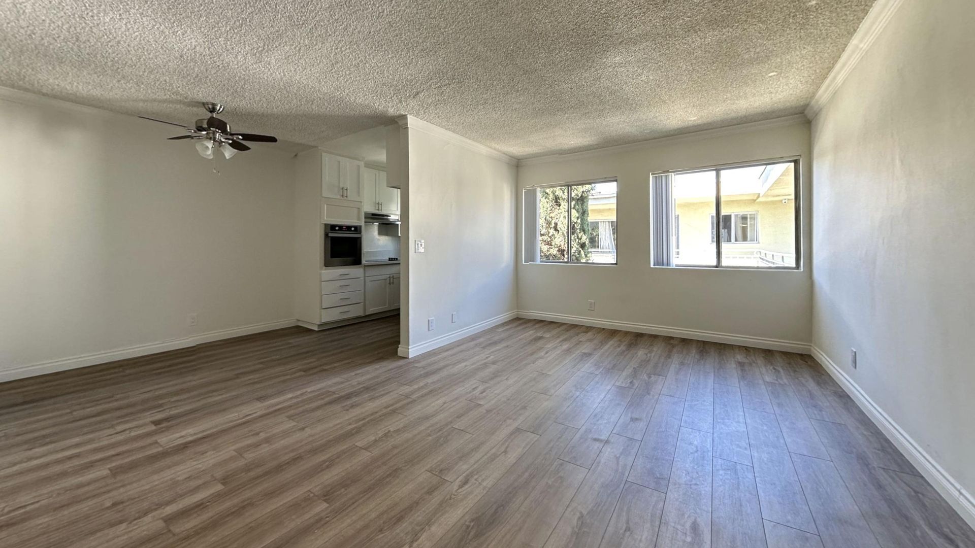 Empty apartment living room with wood-look flooring, two windows, and entry to the kitchen.