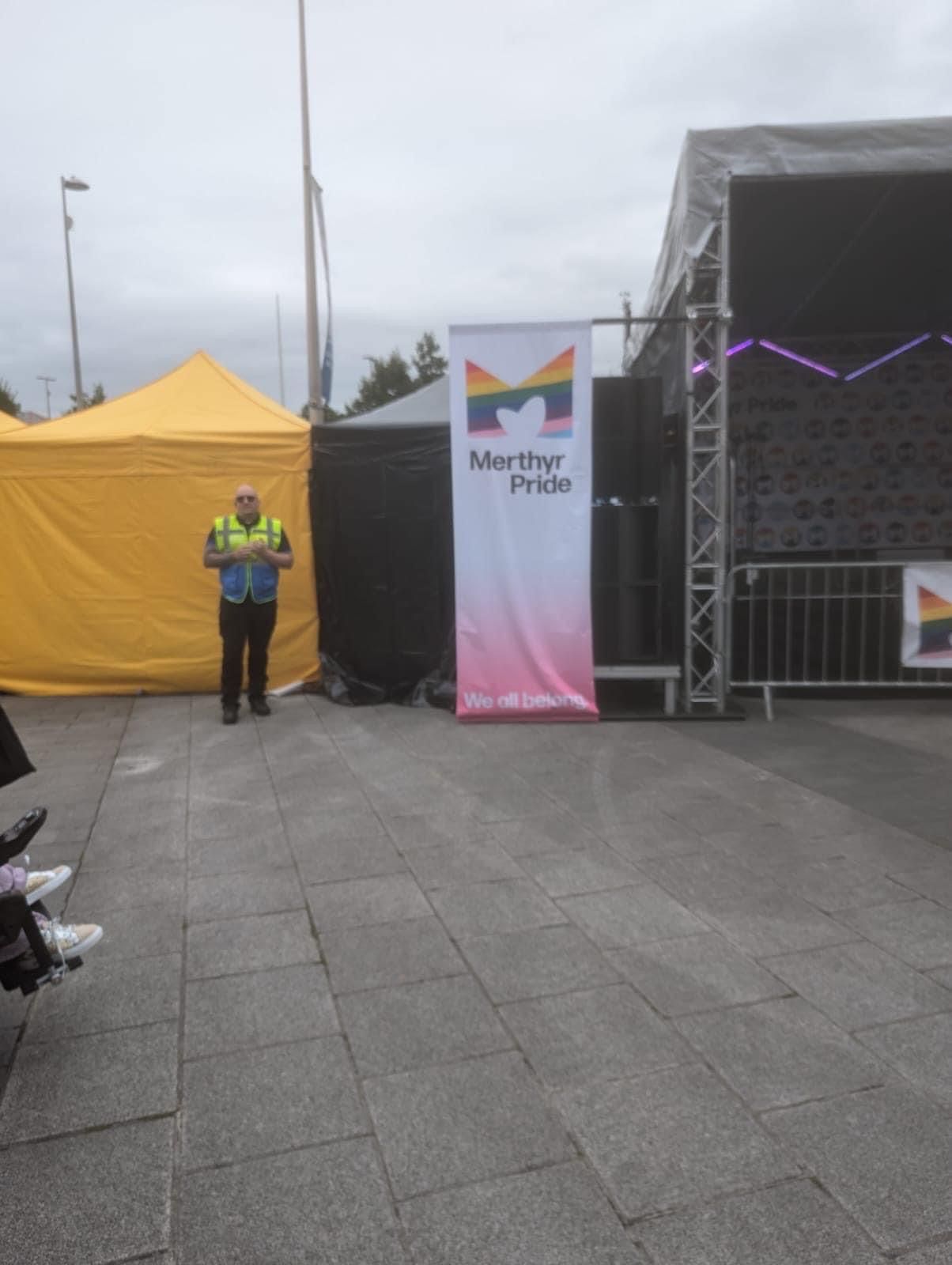 A man in a yellow vest is standing in front of a stage with a banner that says Merthyr pride.