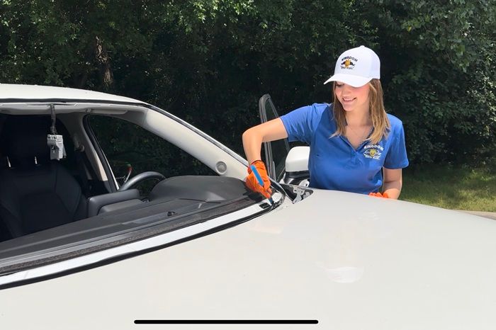 Woman in blue shirt and white cap leaning on a white car outdoors by a wooded roadside
