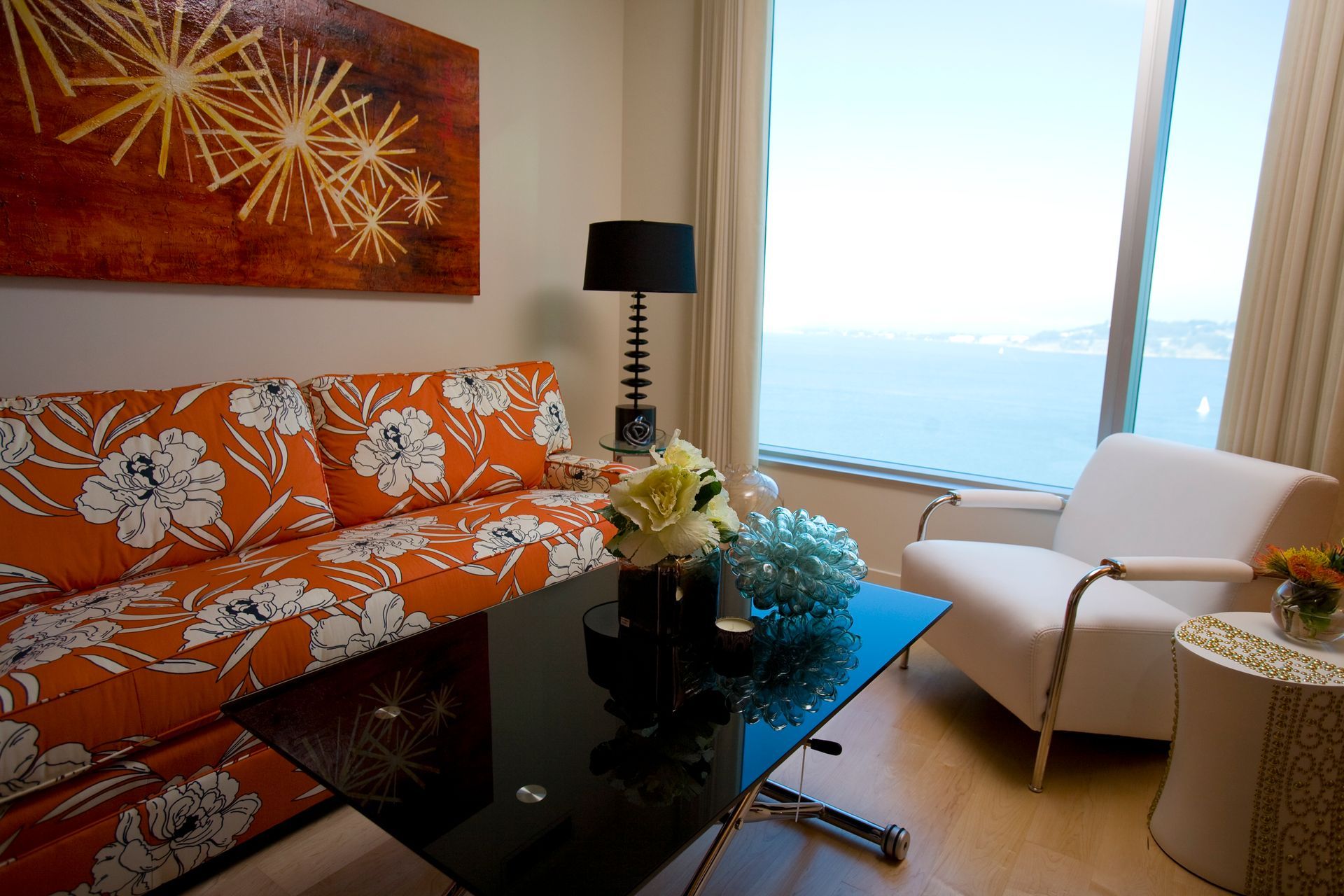 Living room with orange floral couch, black table, white chair, large window with water view.