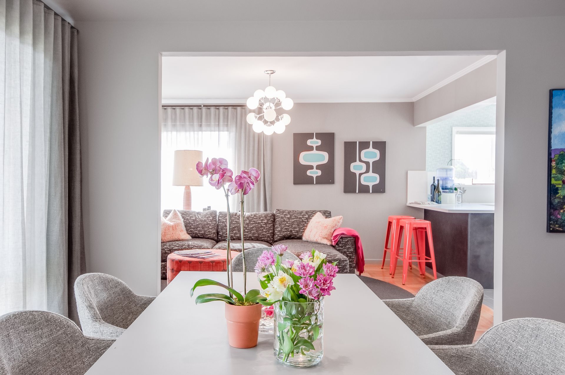 Dining room table with flowers, opening to a living room with sofa, art, and a red stool.