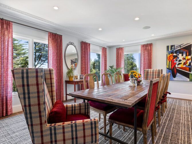 Dining room with a long wooden table and chairs, plaid armchair, red curtains, and colorful artwork.