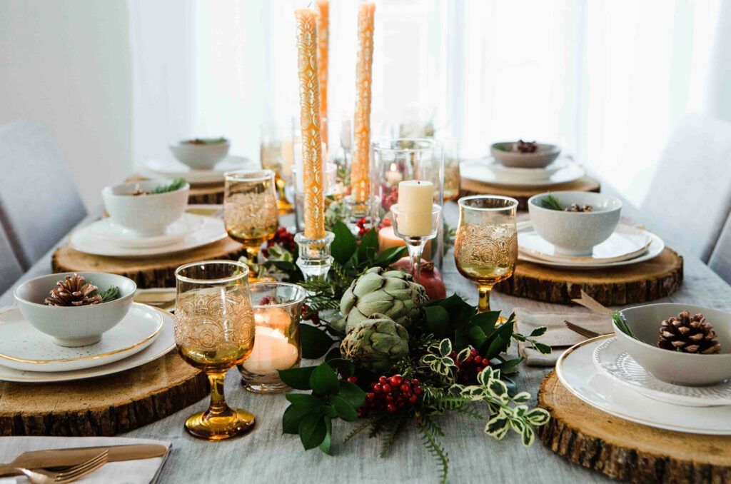 Festive dining table set with gold glassware, white plates, wooden chargers, and a greenery centerpiece.