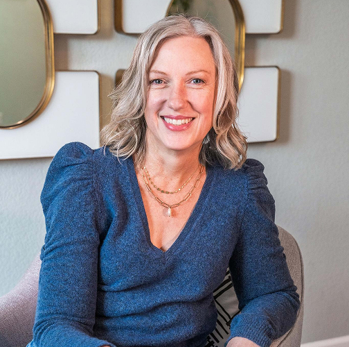 Woman with graying hair smiles, wearing a blue sweater. Sat in a chair. Decorative mirrors on wall behind.