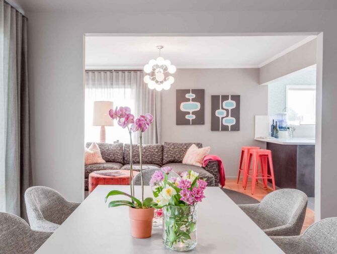 Dining room with table, chairs, flowers, and view of a living room with a sofa, art, and a kitchen.