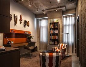 A modern home office with a desk, bookshelf, armchair, and tribal masks on the wall.