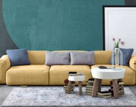Yellow sofa with blue pillows, two small stools on a patterned rug, against a teal wall with a circular design.