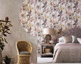 Bedroom with patterned wallpaper, woven chair, bed, lamp, and plants.