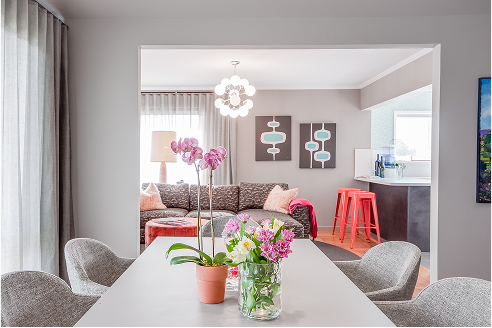 Dining room table with flowers, opening to a living room with sofa, art, and a red stool.