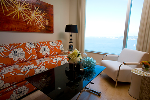 Living room with orange floral couch, black table, white chair, large window with water view.