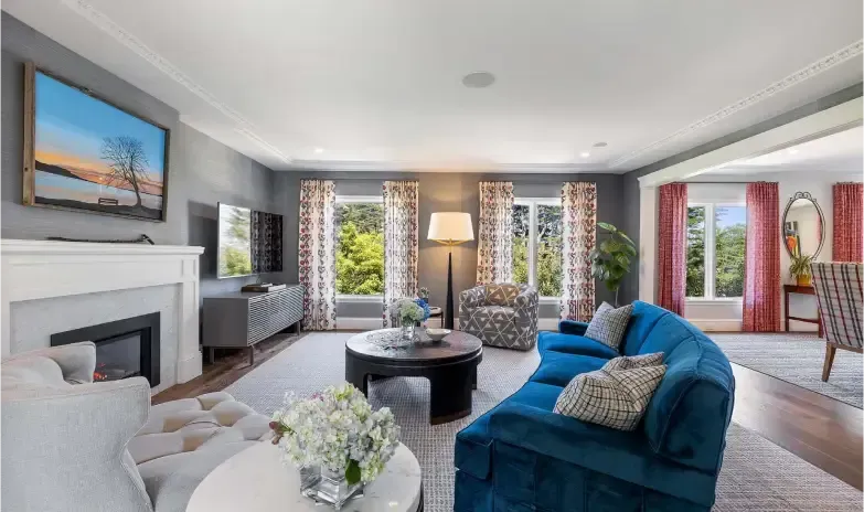 Spacious living room with a fireplace, blue velvet sofa, round coffee table, and large windows with floral curtains.