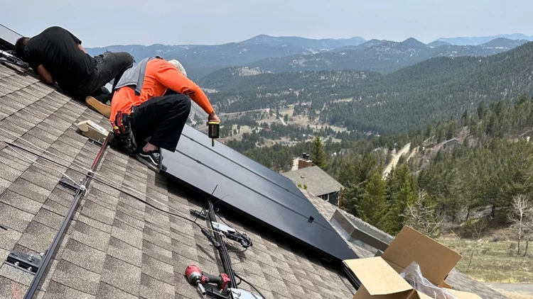 Roofer laying dark gray tiles on a roof. Hands placing tile, boots visible. Blue sky background.
