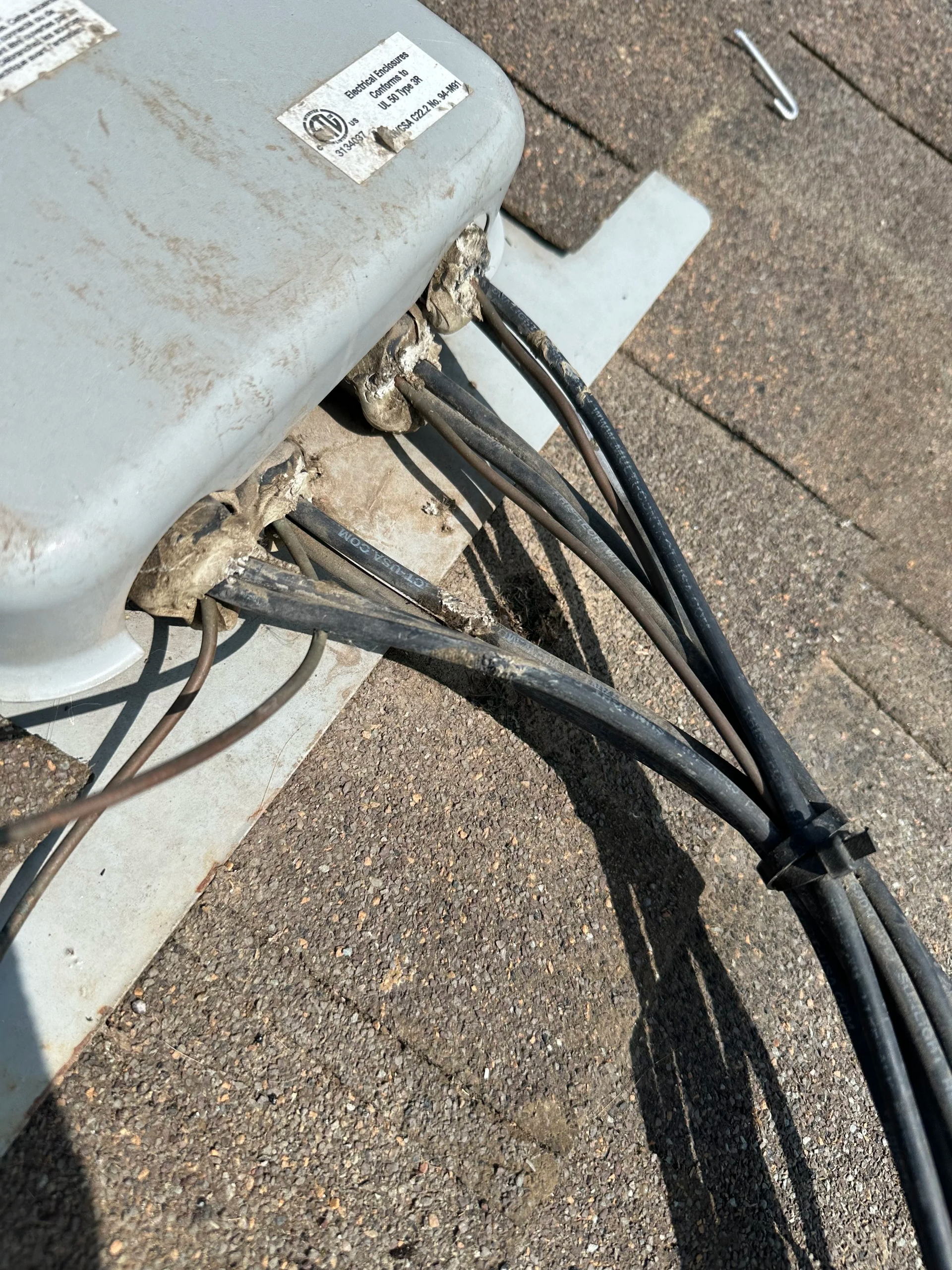 Gray electrical box with multiple black cables emerging, sealed with sealant, on a textured, brown surface.