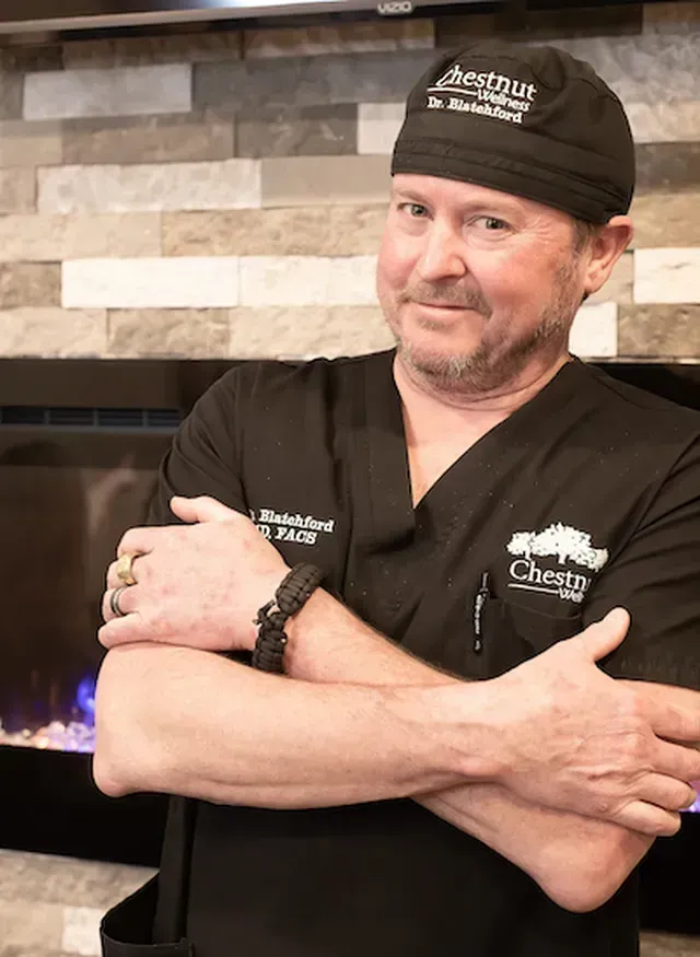 Doctor Blatchford wearing a chestnut wellness hat stands with his arms crossed