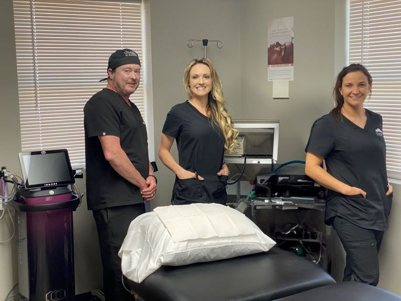 Three medical professionals in scrubs stand near a patient bed with equipment in a clinic room.