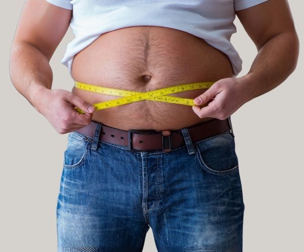 Man in jeans and a white shirt measures his waist with a yellow tape measure.