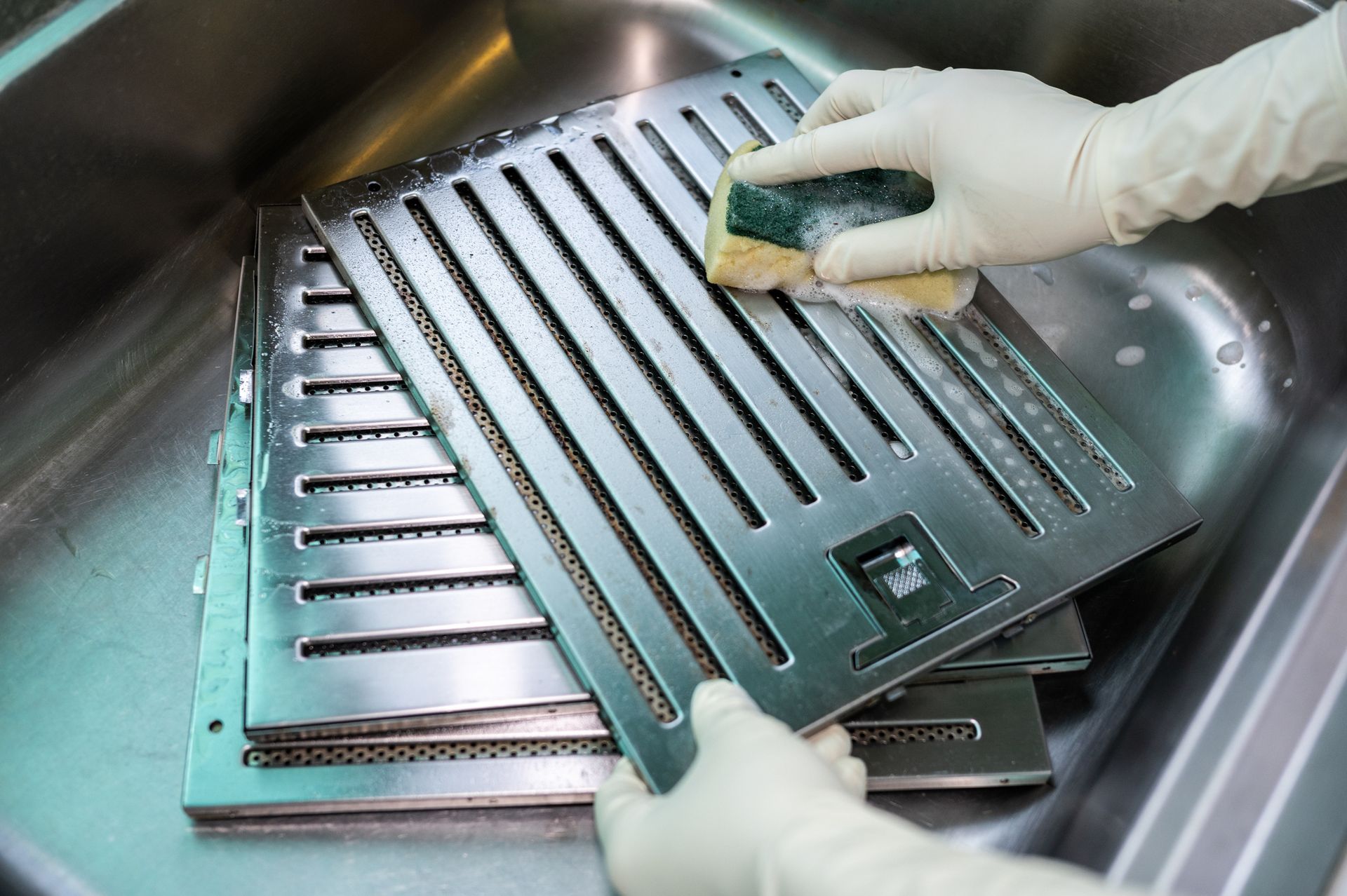 Person wearing gloves scrubbing a metal filter in a sink with a sponge.