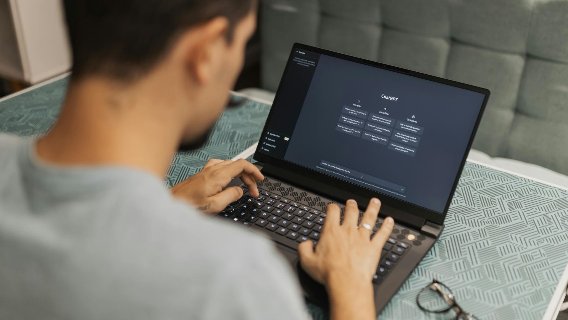 Un homme tape sur un ordinateur portable à écran sombre, posé sur une surface bleue ; ses lunettes sont à proximité.