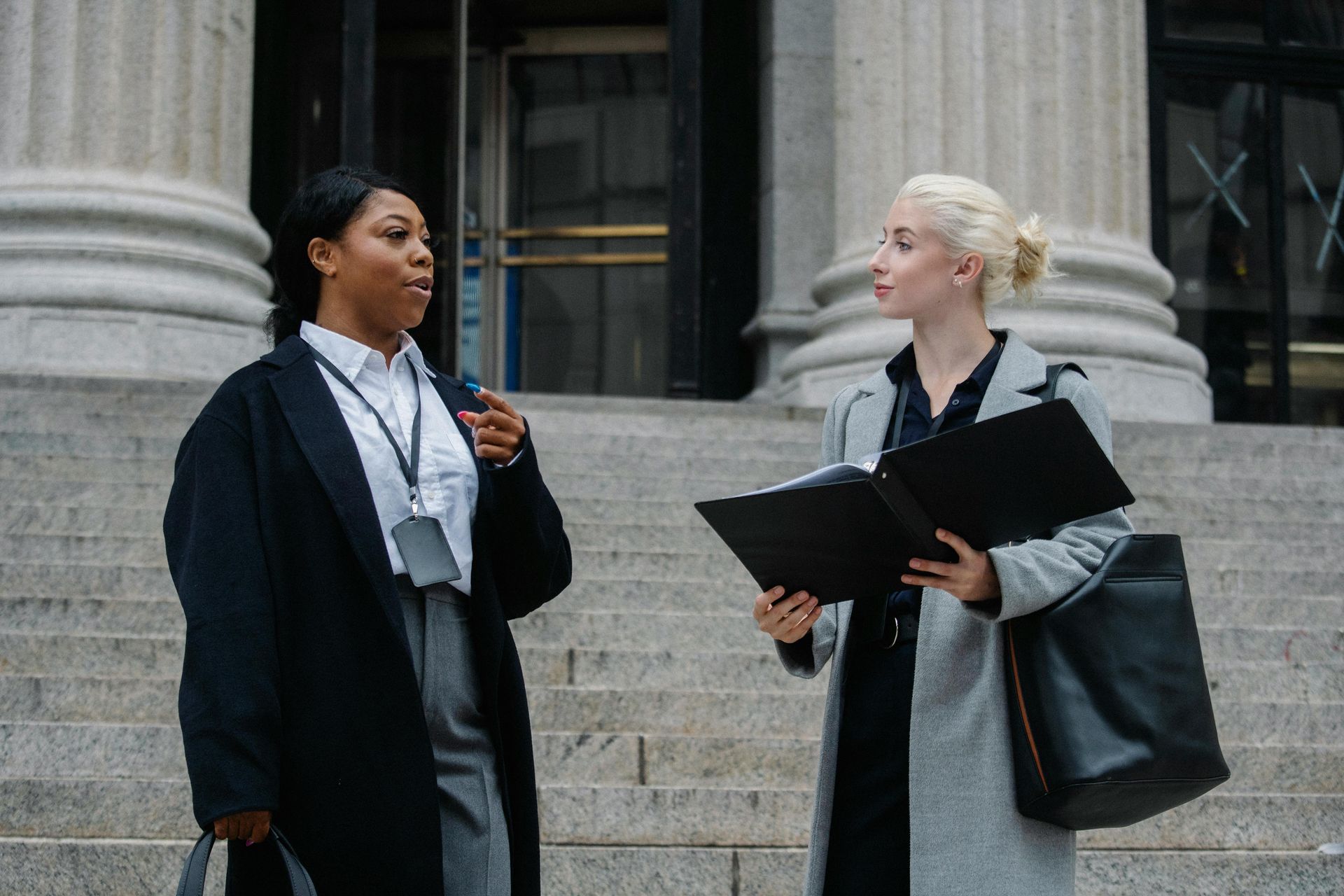 Deux femmes en tailleur devant un palais de justice. L'une parle dans un micro, l'autre tient un classeur.