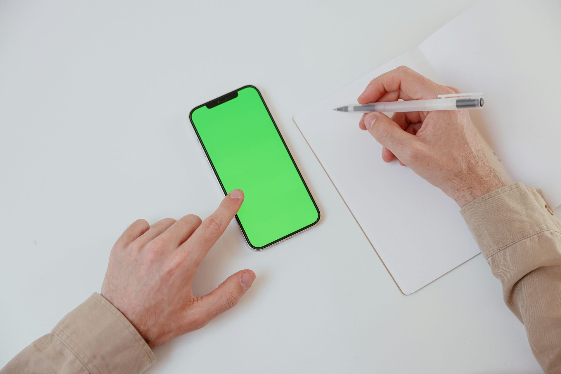 Des mains écrivent dans un cahier et touchent un téléphone à écran vert sur un bureau blanc.