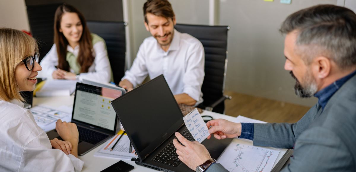Four people in an office setting collaborate at a table with laptops and documents, discussing a project.