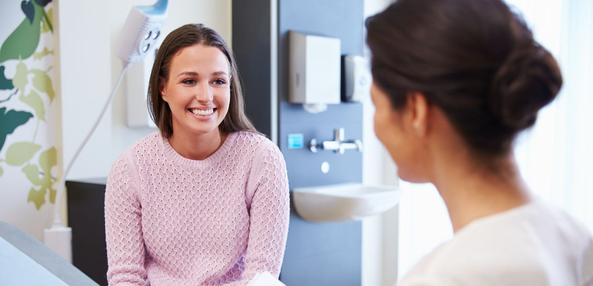 Two women talking in a bright medical office, one smiling on an exam table.